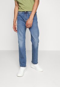 Lichtblauwe spijkerbroek met een rechte pasvorm, ritssluiting en vijf zakken, gecombineerd met witte sneakers en een groene shirt.