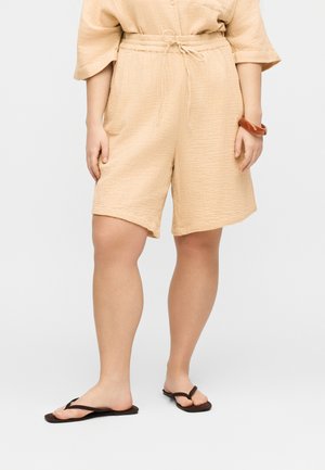 Beige tekstuurilliset shortsit, joissa nyörivyötärö, yhdistetty vastaavaan toppiin, ruskea rannekoru ja mustat varvassandaalit seisovalla henkilöllä.