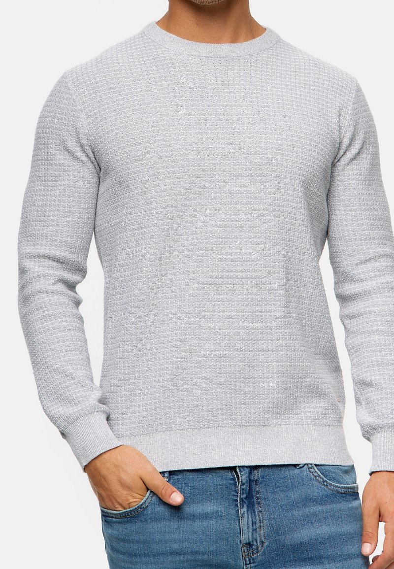 Homme portant un pull gris clair texturé à manches longues et col rond, avec une main dans la poche d'un jean bleu.