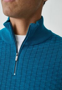 Türkisfarbener Pulli mit Viertelreißverschluss im strukturierten Strickmuster, mit geripptem Kragen und Reißverschluss. Über einem weißen Hemd getragen.