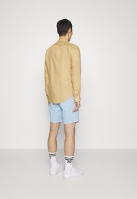 Hnědý lněný dlouhý rukáv, modré šortky, bílé tenisky a pruhované ponožky, viděné z zadní strany na jednobarevném pozadí.