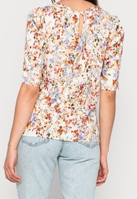 Blommig topp med korta ärmar, vit bas, färgglad blommönster i nyanser av rosa, blått och orange. Har en nyckelhålssvept detalj i ryggen.