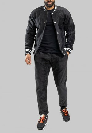 Survêtement en polaire noir avec une veste bomber, des poignets rayés et un pantalon assorti. Porté avec des baskets noires aux accents orange.