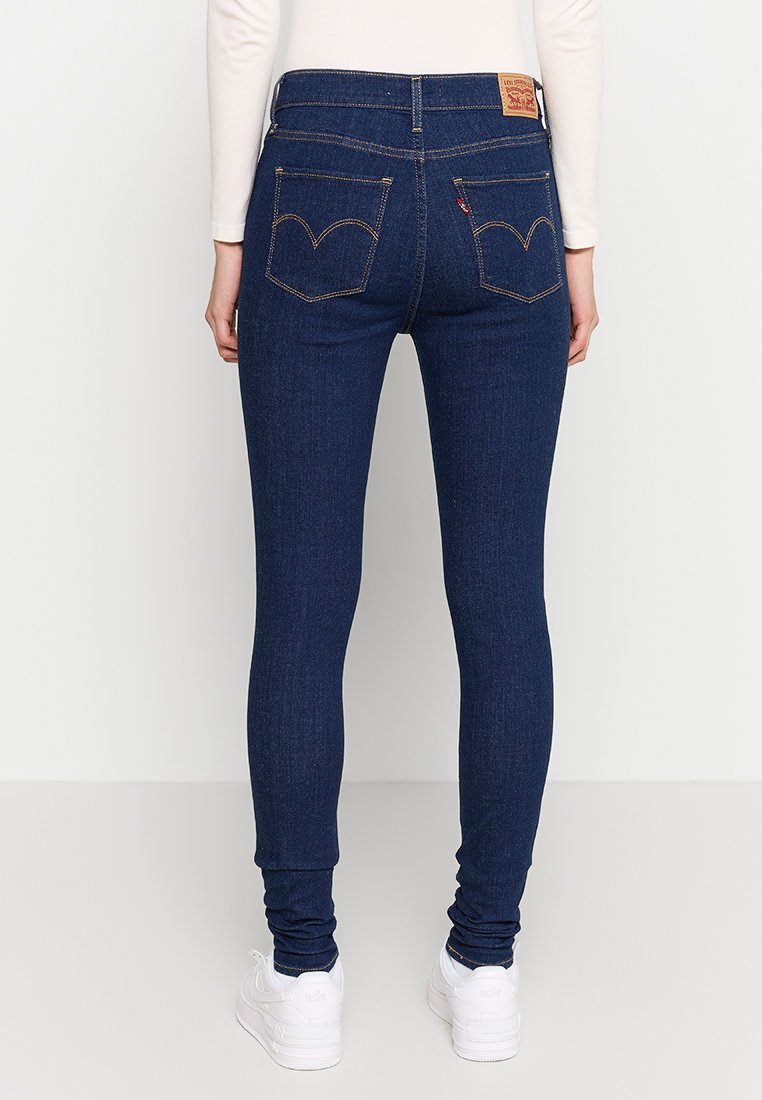 Person in dunkelblauen Skinny Jeans mit Gesäßtaschen, Kontrastnähten, weißen Sneakers und einem weißen Langarmoberteil, steht mit dem Rücken zur Kamera.