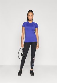 Μωβ αθλητική μπλούζα με κοντά μανίκια και εφαρμοστή σχεδίαση. Μαύρα κολάν με την ένδειξη "ACTIVE" εκτυπωμένη σε κλίμακα χρωμάτων, συνδυασμένα με μαύρα αθλητικά παπούτσια.