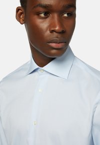 Světle modrá košile s klasickým límcem, předním zapínáním na knoflíky a hladkou texturou látky. Knoflíky jsou bílé s mírným leskem.