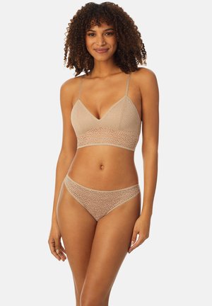 Kobieta o kręconych włosach, ubrana w beżowy koronkowy biustonosz i dopasowane koronkowe majtki, stojąca i uśmiechająca się na białym tle.