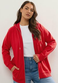 Sudadera roja con cremallera y capucha ajustable, mangas largas y bolsillos tipo canguro. Combinada con una camiseta blanca y jeans de tiro alto azules.