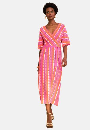 Žena oblečená v ružovom, oranžovom a bielom pruhovanom zavinovacom šate s krátkymi rukávmi a béžových platformových sandáloch stojaca pred jednofarebným pozadím.