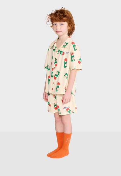 Enfant aux cheveux roux bouclés portant un ensemble de pyjama crème avec des motifs verts et orange et des chaussettes orange vif, debout devant un fond uni.