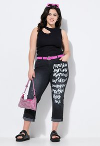 Top negro sin mangas con un corte, combinado con jeans negros desgastados que tienen texto blanco, cinturón rosa y bolso rosa brillante. Sandalias de plataforma negras.