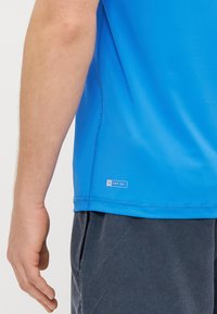 Zils UPF 50+ krekls ar īsām piedurknēm un plakano šuvēm; apakšējā apmalē ir mazs uzlīmēm. Audums izskatās gluds un viegls.