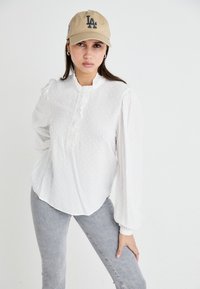Camicia bianca con pois, colletto arricciato e chiusura a bottoni. Abbinata a jeans grigi e un cappellino beige con il logo "LA".