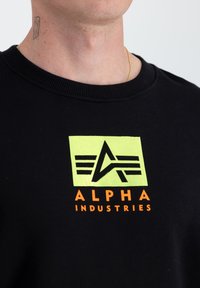 Fekete pulóver, kerek nyakkivágással, elől élénk zöld és narancssárga "ALPHA INDUSTRIES" logóval és grafikai designnal.