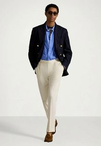 Marineblå dobbeltradet blazer, lyseblå skjorte, beige bukser, brune loafers og solbriller brukt av en mann som står foran en lys bakgrunn.