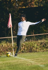 Atleet in een wit en zwart shirt met lange mouwen, groene broek en zwarte voetbalschoenen, die zich voorbereid om een gele voetbal te schoppen nabij een roze hoekvlag.