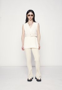 Top sem mangas de malha branca com um cinto largo, combinado com calças de creme com nervuras e sandálias chunky pretas com detalhes brancos.