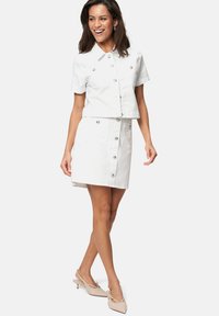 Chemise blanche à manches courtes, coupée au niveau du ventre, associée à une jupe blanche avec des boutons en argent et des poches avant.