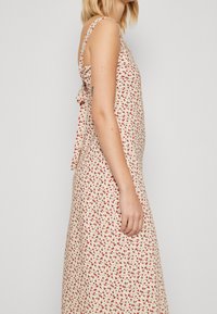 Vestido floral hecho de una tela ligera. Presenta flores rojas sobre un fondo crema, con tirantes y un detalle de lazo en la espalda.