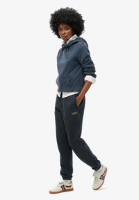 Marineblauwe hoodie met trekkoord, voorzak en logodetail. Gepaard met bijpassende sportbroeken en witte sneakers met rode accenten.