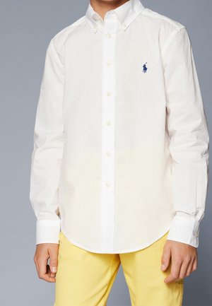 Dziecko ubrane w białą, długorękawową koszulę z kołnierzykiem i żółte spodnie, stojące na jednolitym tle.