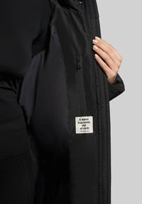 Sort jakke med glat stof indvendig, der har en sidelomme med lynlås og et hvidt mærke med tekst. Hånd berører stoffet.