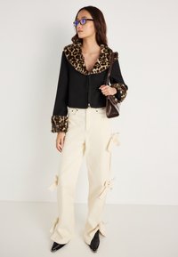 Jaqueta preta curta com colarinho e punhos de pelo de leopardo; combinada com calças off-white com laços laterais e sapatos pontiagudos pretos.