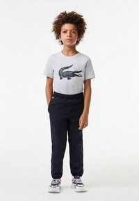 Grå bomulls-T-skjorte med en mørk krokodillegrafikk, kombinert med mørk marineblå bukser med elastisk midje. Hvite og marineblå joggesko med røde detaljer.