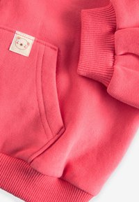 Rožinės spalvos džemperio rankovė ir ribotu sagtimi su priekinės kišenės mažu smėlio spalvos lopu, kuriame pavaizduota paprasta meškos veido iliustracija.