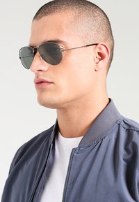 Ochelari de soare cu lentile gri și rame din metal. Modelul poartă o jachetă bomber gri peste o cămașă albă, prezentând o vedere detaliată din profil.
