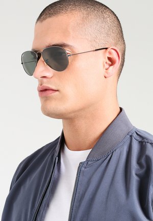Occhiali da sole con lenti grigie e montatura in metallo. Il modello indossa una giacca bomber grigia sopra una maglietta bianca, mostrando una vista ravvicinata di profilo.