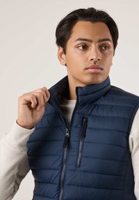 Giovane uomo che sistema il colletto di un gilet trapuntato blu navy indossato sopra una camicia beige a maniche lunghe, guardando di lato.