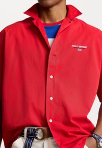 Red button-up shirt with a sharp collar and white logo detailing. Features smooth texture, casual fit, paired with a striped belt.