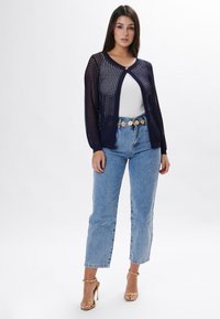 Navy-blauer Strickcardigan mit V-Ausschnitt und offenem Vorderteil, ausgestattet mit einer Knopfleiste, kombiniert mit hellblauen Jeans und goldenen Riemchenschuhen.