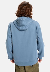 Modrá mikina s kapucí vyrobená z měkké tkaniny, s prostornou kapucí, ribovými manžetami a klokankou. Zadní pohled ukazuje volný střih.