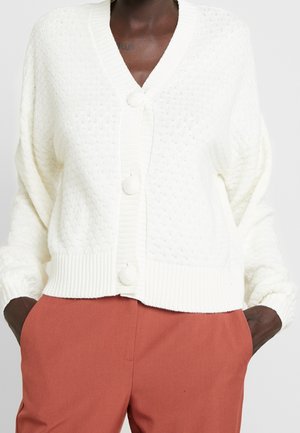 Personne à la peau foncée portant un cardigan en tricot blanc texturé avec de gros boutons et un pantalon couleur rouille, les mains dans les poches.