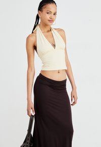 Crème-kleurige haltertop met een diepe V-hals en plooien; aansluitende zwarte maxi-rok; model houdt een donkere handtas vast. Soepele, rekbare stof.