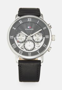Reloj con correa de cuero negra y caja de tono plateado, esfera negra con números romanos, subesferas y un logotipo. Diseño de fondo texturizado.