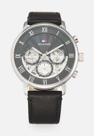 Montre à bracelet en cuir noir avec boîtier en métal argenté, cadran noir présentant des chiffres romains, des sous-cadrans et un logo. Design de fond texturé.