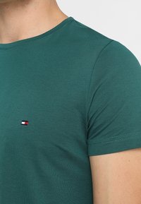 Tummanvihreä lyhythihainen t-paita pehmeästä kankaasta, jossa on pieni brodeerattu logo punaisilla, valkoisilla ja sinisillä väreillä rinnassa.
