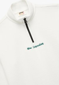Sudadera blanca con cremallera, de superficie texturizada, que presenta el texto verde bordado "Nueva Generación" en el frente y una cremallera negra.