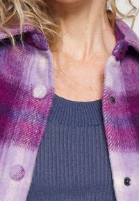 Fialová a růžová kostkovaná vlněná košile s bundou s kulatými knoflíky, nošená přes tmavě modrý ribbový svetr. Texturovaná látka, ležérní design.