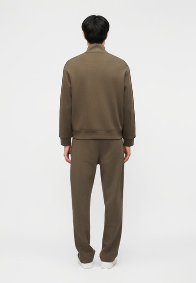 Άτομο που φοράει καφέ σετ φόρμας, στέκεται με την πλάτη γυρισμένη προς ένα απλό φωτεινό φόντο.