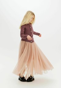 Cárdigan de punto morado con botones redondos, superpuesto a una falda larga de tul ligera y transparente en tono rosa claro con acentos de lunares, acompañada de zapatos negros brillantes.