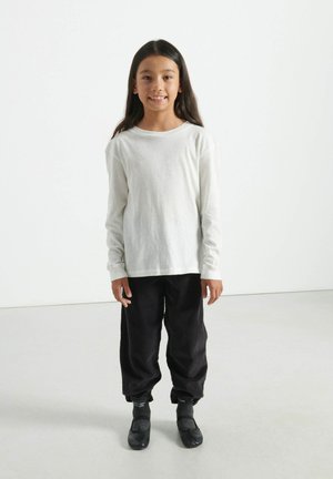 Camisa blanca de manga larga, pantalones amplios negros con textura, calcetines oscuros y zapatos planos negros; de pie sobre un suelo claro contra un fondo blanco.