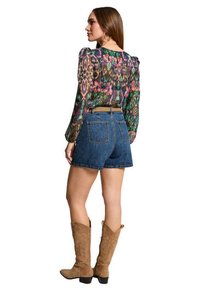 Blouse à motifs multicolores avec des manches bouffantes, associée à un short en denim taille haute et des bottes de cow-boy beiges. Vue de dos.