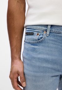 Světlemodré džíny s úzkým střihem, s měděnými akcenty a černou nášivkou Calvin Klein na zadní kapse.