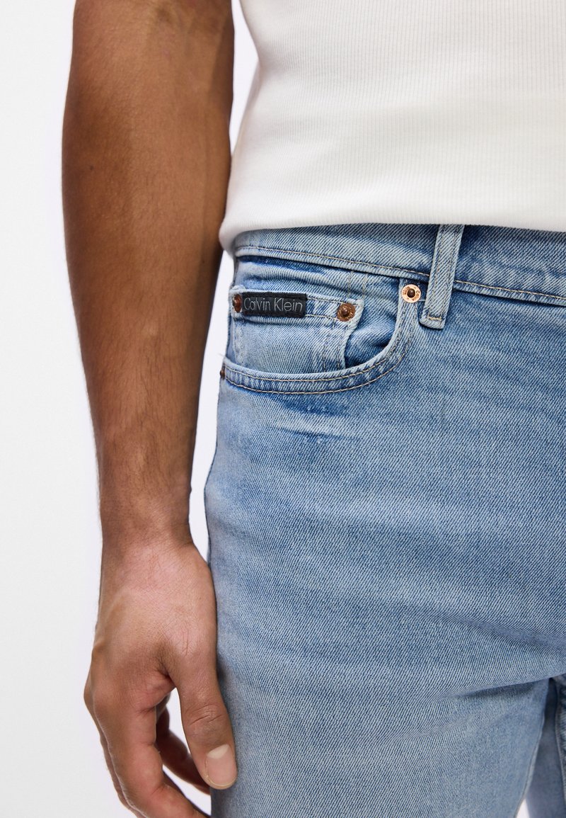 Light blue denim jeans with a slim fit, featuring copper accents and a black Calvin Klein label on the back pocket.