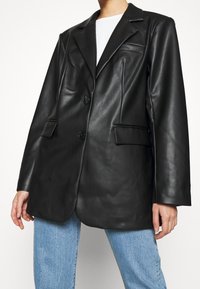 Sort læderblazer med en skræddersyet pasform, enknapsdesign, to forlommer og en glat overflade. Båret over lyseblå jeans.