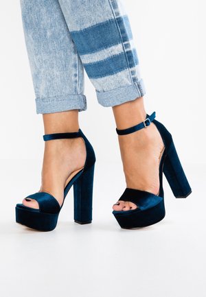 Fødder iført mørkeblå fløjl platform høje hæle med ankelstropper kombineret med opkrøllede lyseblå jeans med hvide striber.